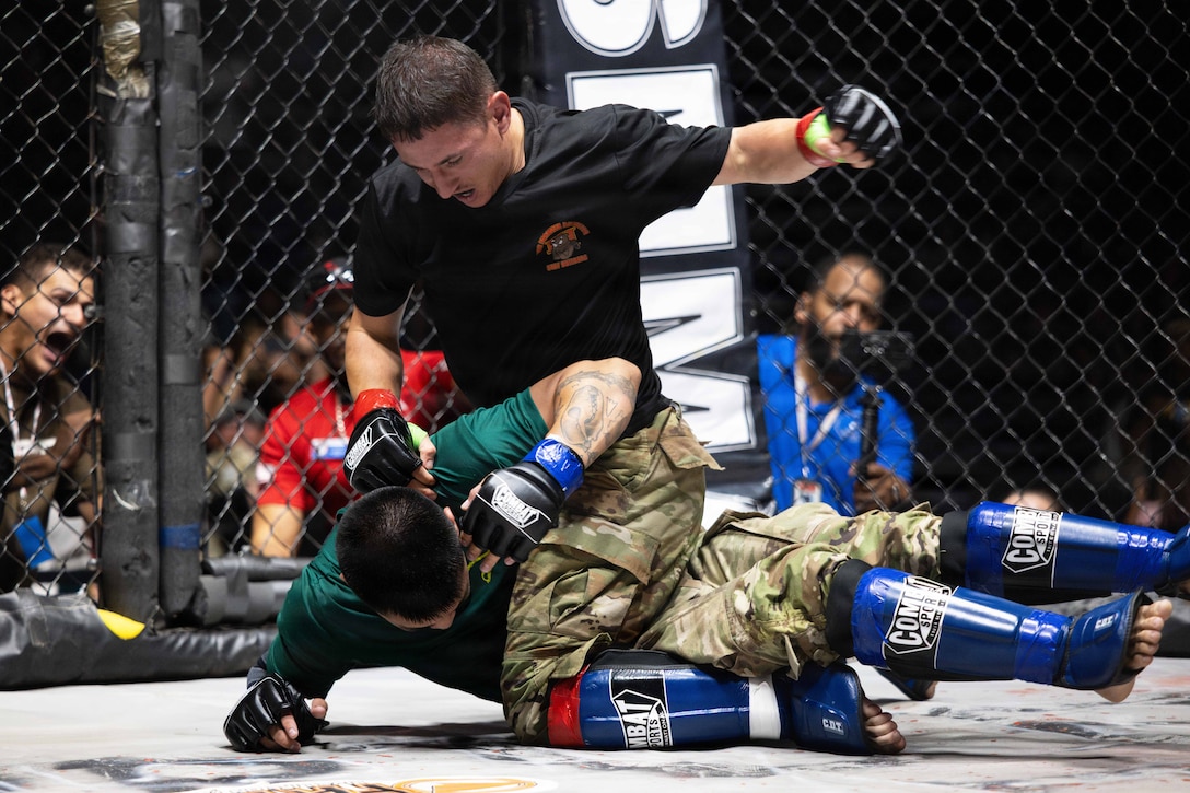 A soldier in fighting gear sits on another soldier while preparing to punch them as spectators watch through a fence.