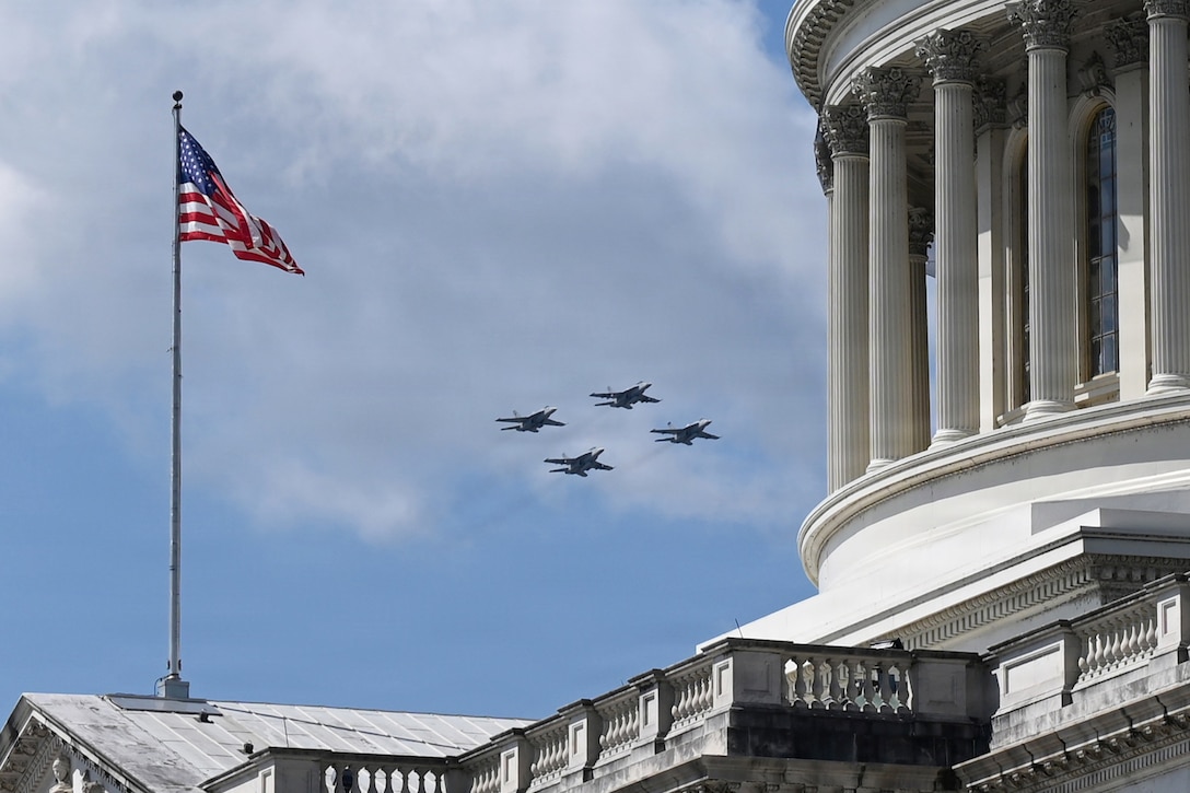 Four aircraft fly in formation in a blue sky toward a white monumental building as an American flag waves to the left.