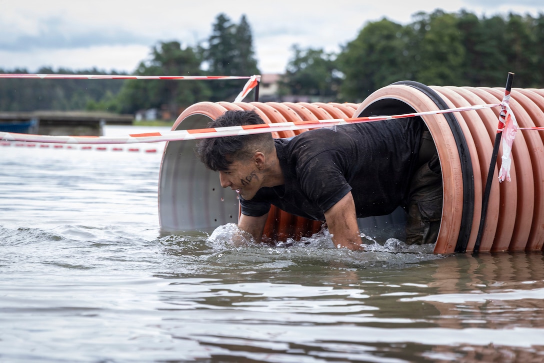 A soldier climbs out of an orange tunnel into low murky waters in between roped barriers with trees in the distance.