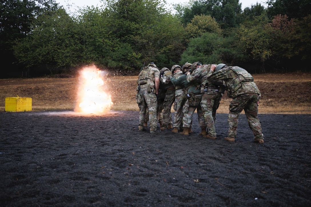 Soldiers take cover behind one another's backs while standing in formation in the dirt as an orange explosion is visible to the left on a gloomy day.