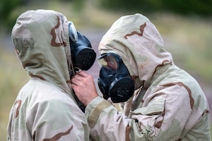 A person wearing a camouflage military uniform and gas mask helps another person in similar attire put their face mask on.