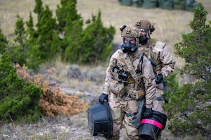 Two people wearing camouflage military uniforms, tactical gear and gas masks transfer equipment to a training site in the woods.