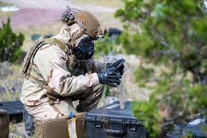 A person wearing a camouflage military uniform, tactical gear and gas mask kneels in the woods while looking at a tablet.