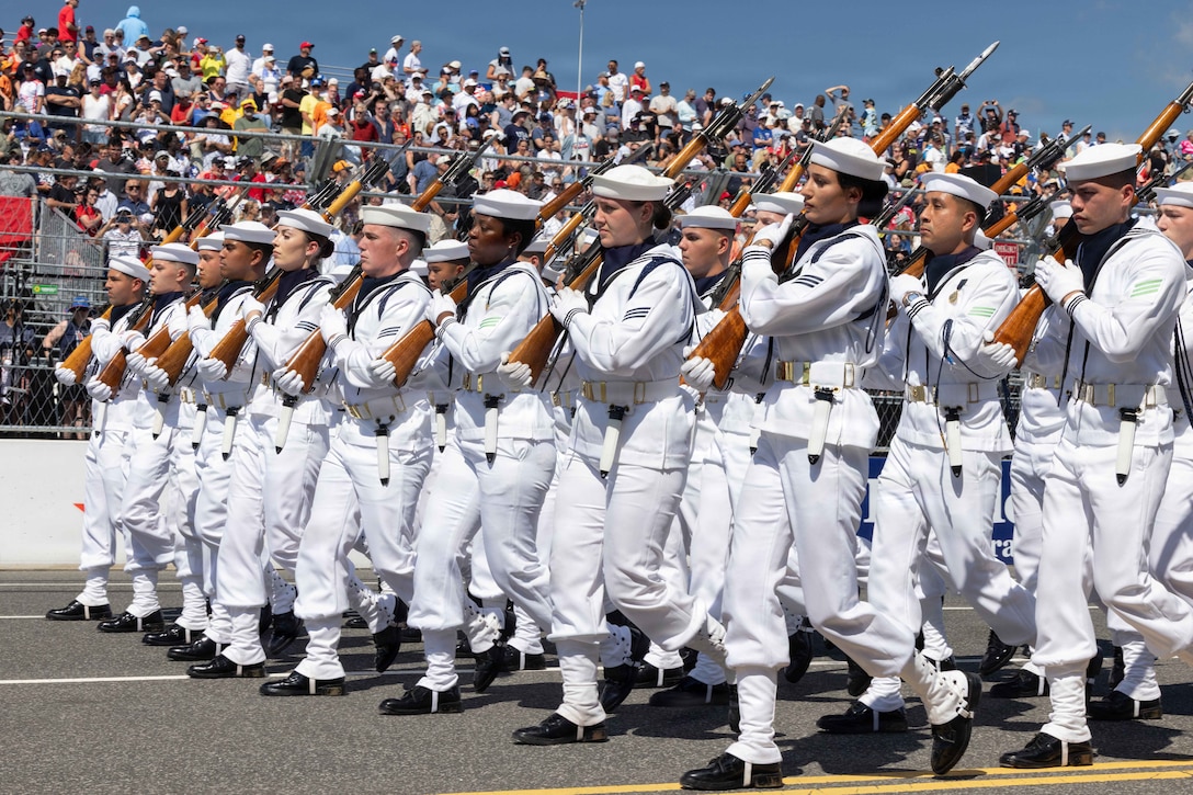 Sailors in white uniforms and sailor caps hold rifles fitted with bayonets over their right shoulders and march in formation along a racetrack on a sunny day as people watch from bleachers.
