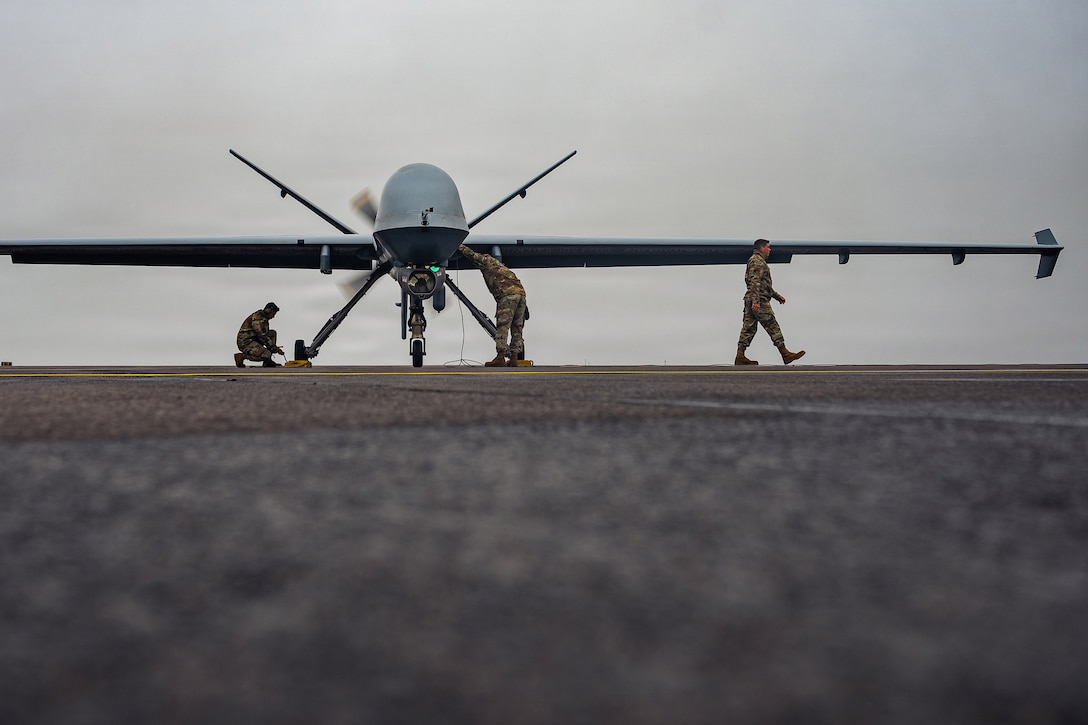 Two airmen work on a large drone parked on a tarmac on a gray day, and another airman walks away.