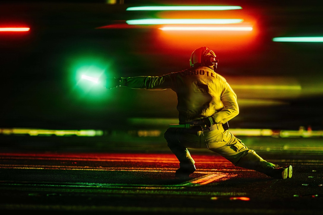 A sailor on a flight deck at night lunges to the left while holding a green light in their left hand. Streaks of green and red lights add color to the darkness.