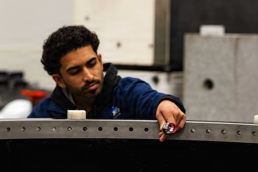 A man in casual attire holds a tool against a large, curved metal device.