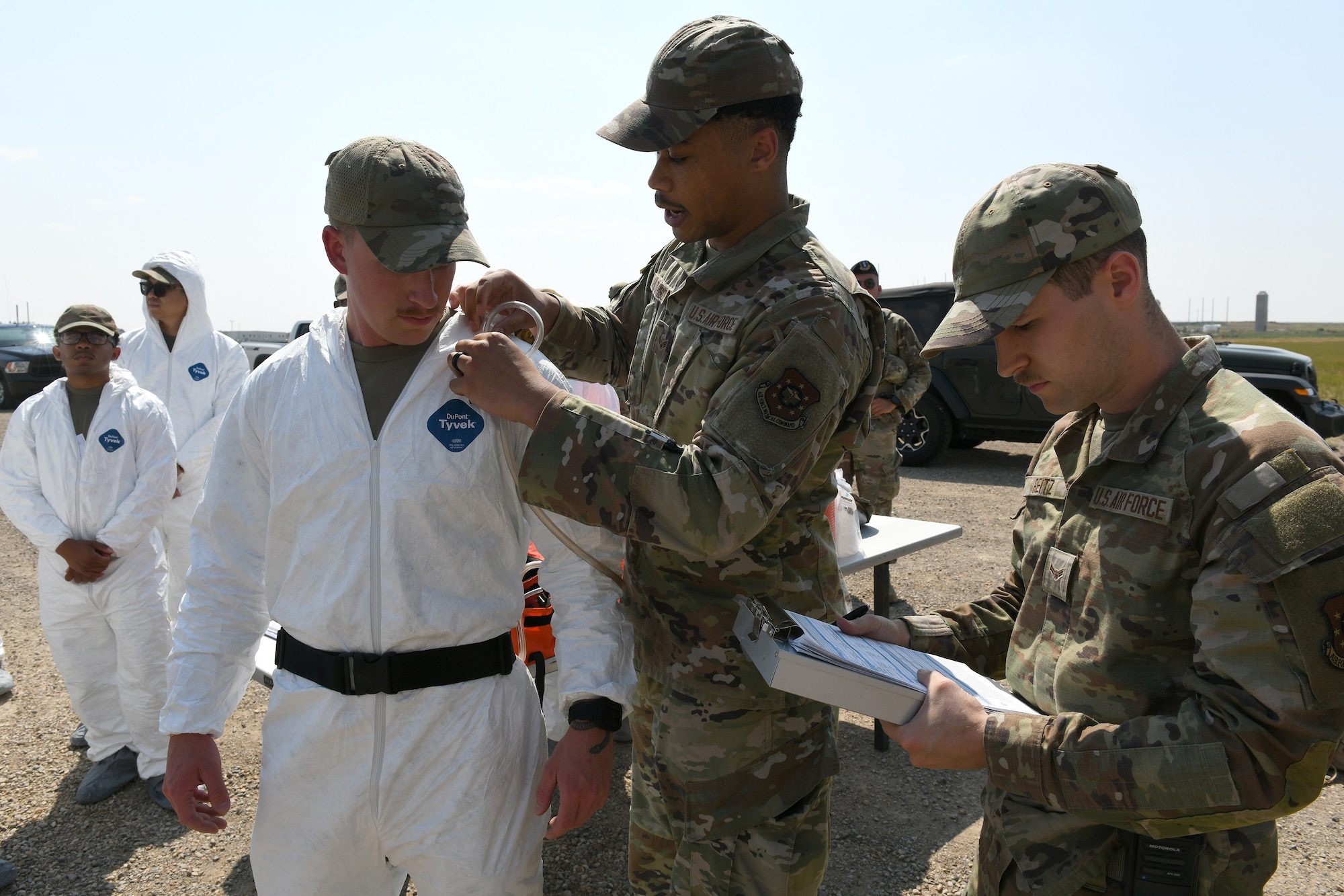 Bioenvironmental Airmen with Malmstrom AFB affix an air quality detection device to an Airman in a white safety suit during a Mass Accident Response Exercise at Malmstrom AFB.
