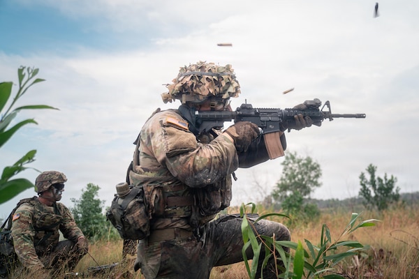 A man in a camouflage military uniform and tactical gear fires his rifle in a jungle clearing, as another man in similar attire observes.