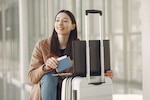 Woman in a brown coat and jeans sits next to her luggage while holding a passport