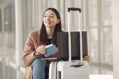 Woman in a brown coat and jeans sits next to her luggage while holding a passport