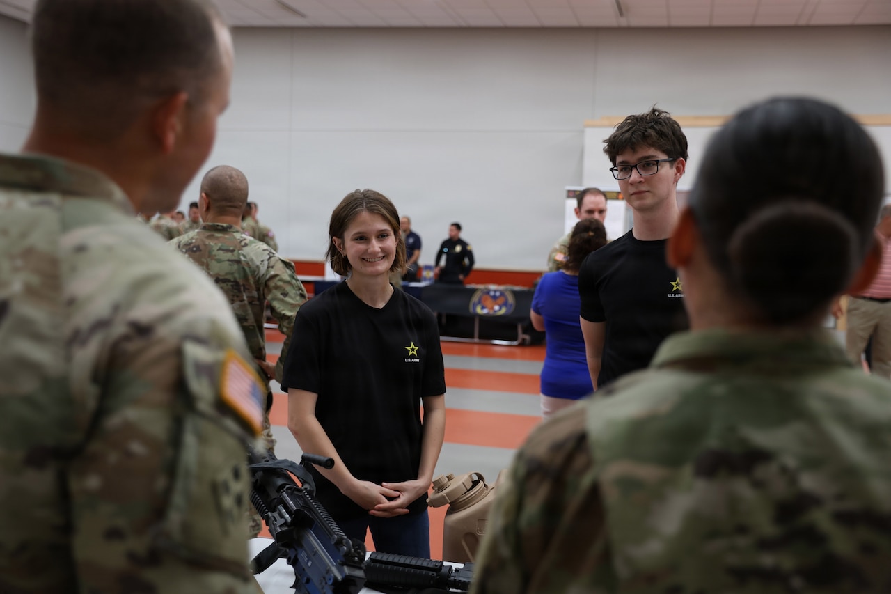 A woman and a man in casual attire speak to two people in camouflage military uniforms.