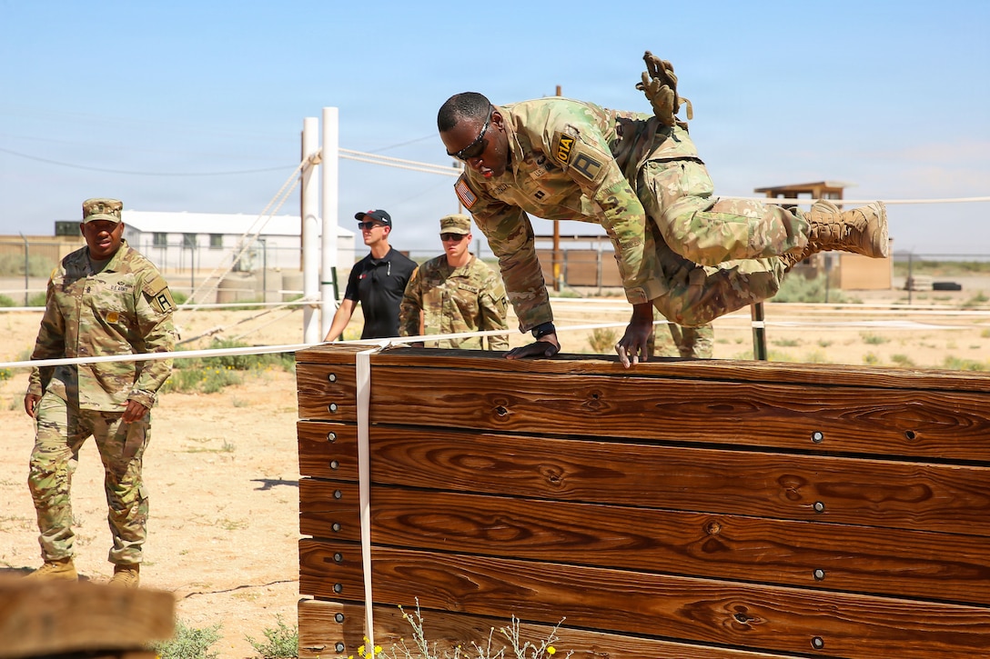A soldier in uniform jumps over a wooded structure in a desert-like area as fellow soldiers watch during the day.