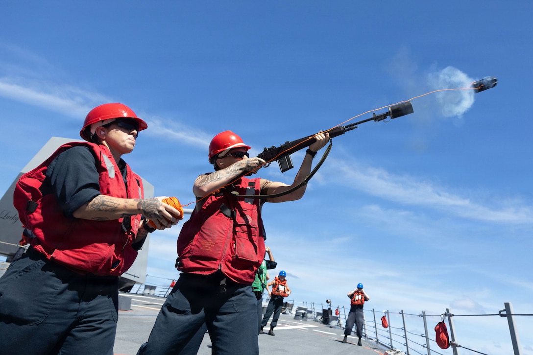 A sailor fires a shot line next to another sailor holding the line aboard a ship at sea during the day.
