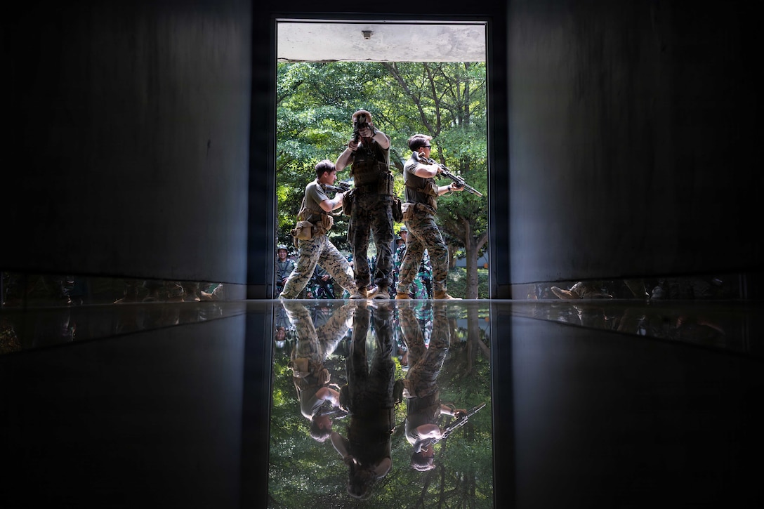 Three U.S. and Indonesian marines stand in a trio formation while holding weapons in the open doorway of a dark room with daylight and trees in the background.