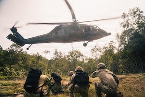 Five people in camouflage military uniforms kneel in an open area of the jungle as a military helicopter hovers overhead.
