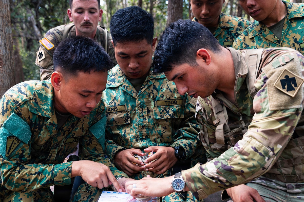 Three people in camouflage military uniforms coordinate a location on a map while three other people in similar attire stand behind them in the jungle.