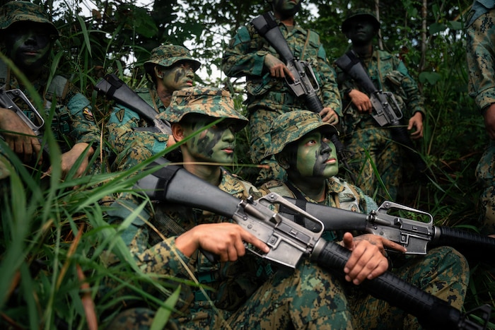 Six people in camouflage military uniforms and face paint gather in the jungle while holding rifles.