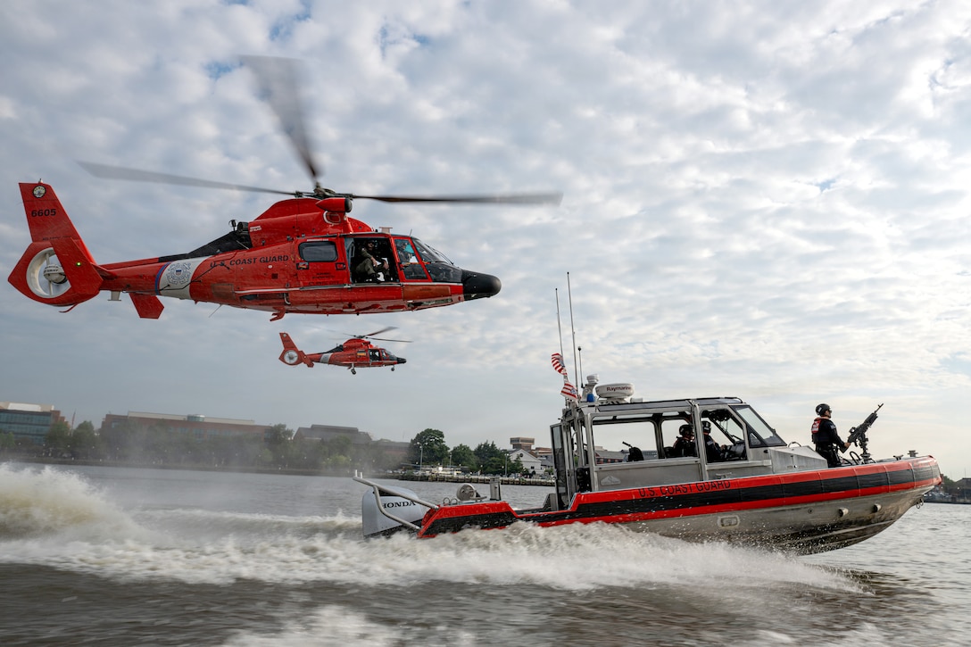 Two orange helicopters fly low over a body of water, following a boat with orange gunwales and a Coast Guardsman standing in the bow manning a mounted machine gun.