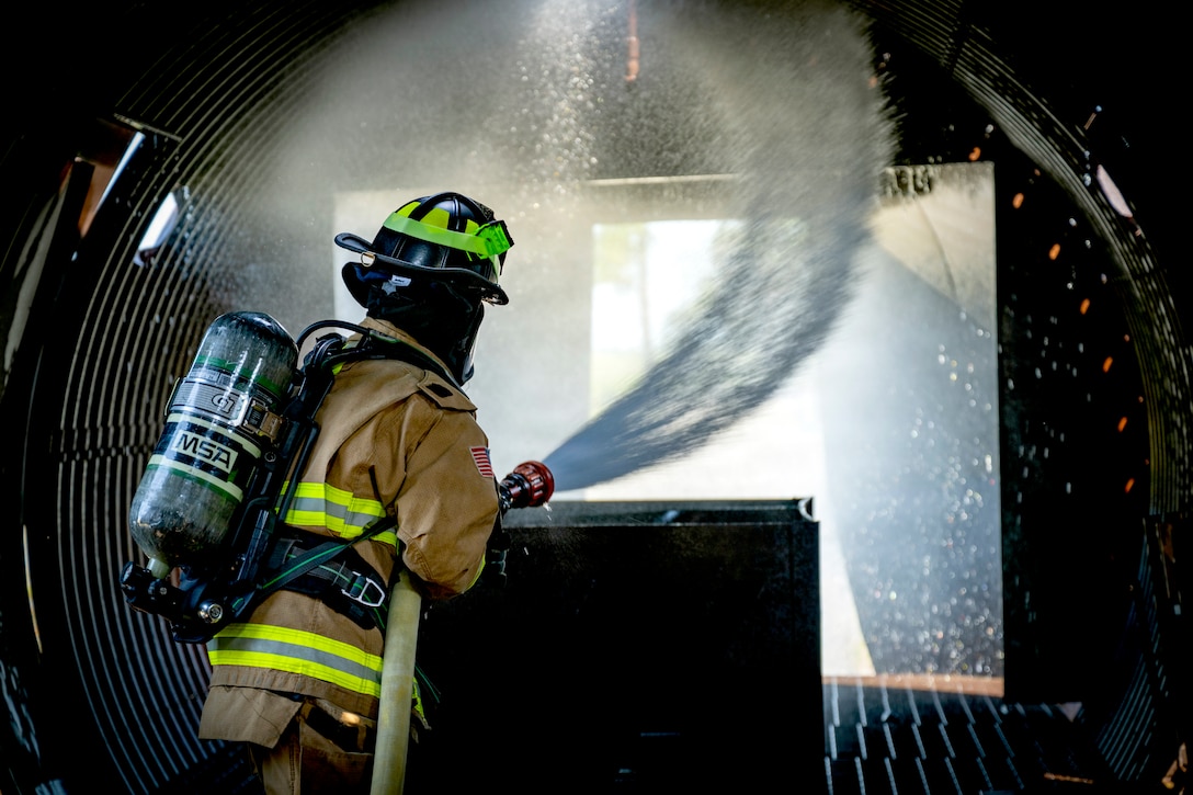 An airman wearing firefighting gear sprays a fire hose inside a building.