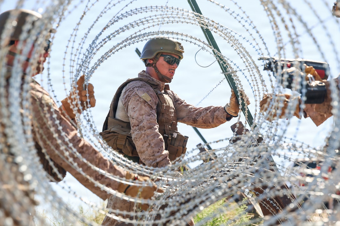 Three Marines wearing camouflage uniforms, helmets and tactical vests uncoil concertina wire and affix it onto green supports outdoors during daytime.