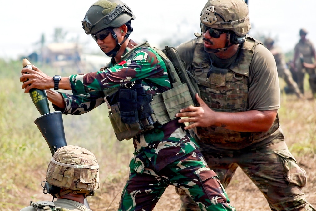 A soldier wearing a camouflage uniform, helmet and tactical vest drops a round into a mortar tube as another soldier wearing a different shade of camouflage uniform and similar gear stands behind him and another one kneels next to the mortar tube.