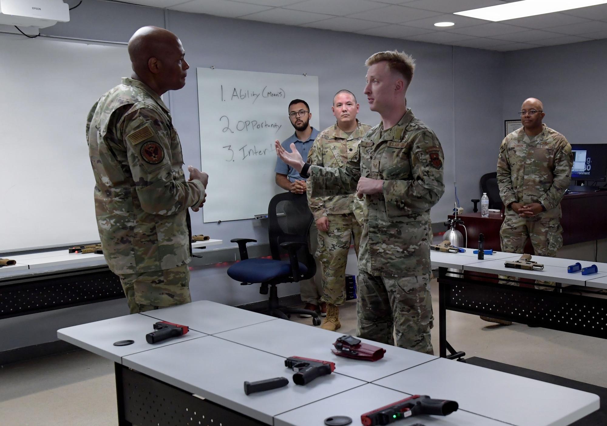 A man wearing a U.S. Air Force uniform speaks to another Airmen inside a training classroom.