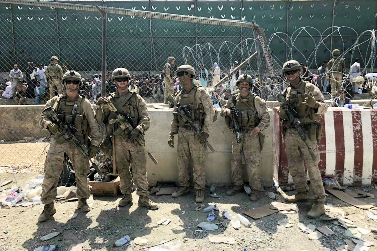 Five men wearing camouflage military uniforms and tactical gear stand outside in front of a concrete vehicle barrier posing for a photo. People in civilian attire are sitting in the background.