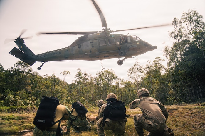 Five people in camouflage military uniforms kneel in an open area of the jungle as a military helicopter hovers overhead.
