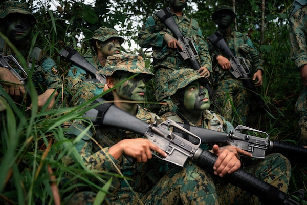Six people in camouflage military uniforms and face paint gather in the jungle while holding rifles.