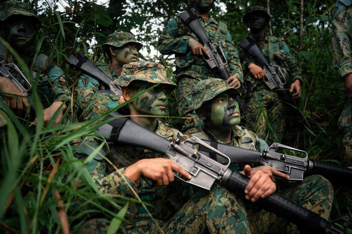 Six people in camouflage military uniforms and face paint gather in the jungle while holding rifles.