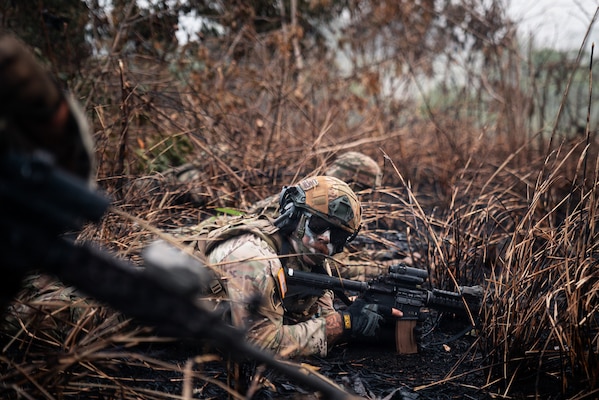 A man in a camouflage military uniform and tactical gear lies down in mud and weeds with his rifle.