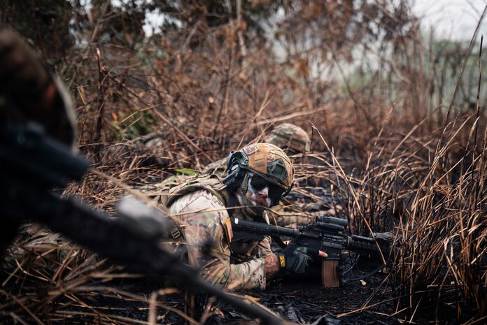 A man in a camouflage military uniform and tactical gear lies down in mud and weeds with his rifle.