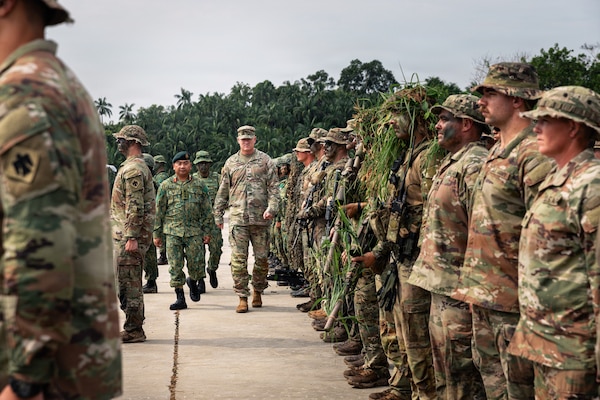 Two men in camouflage military uniforms walk between dozens of other people in similar attire standing in formation outside; there are trees in the background.