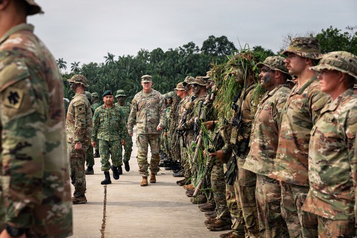 Two men in camouflage military uniforms walk between dozens of other people in similar attire standing in formation outside; there are trees in the background.