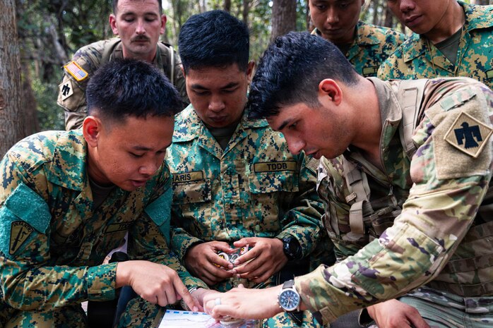 Three people in camouflage military uniforms coordinate a location on a map while three other people in similar attire stand behind them in the jungle.
