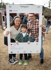 A family, made up of a mom, dad and a toddler boy pose for a photo, holding an Instagram frame sign