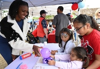 A woman hands a purple balloon to a group of 3 children, 1 toddler age, 2 elementary school age, across a white table. she smiles at them.
