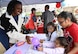 A woman hands a purple balloon to a group of 3 children, 1 toddler age, 2 elementary school age, across a white table. she smiles at them.