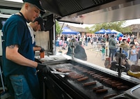 A man in a navy blue shirt uses tongs to flip hot dogs on a flat top grill inside of a food truck