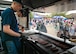 A man in a navy blue shirt uses tongs to flip hot dogs on a flat top grill inside of a food truck