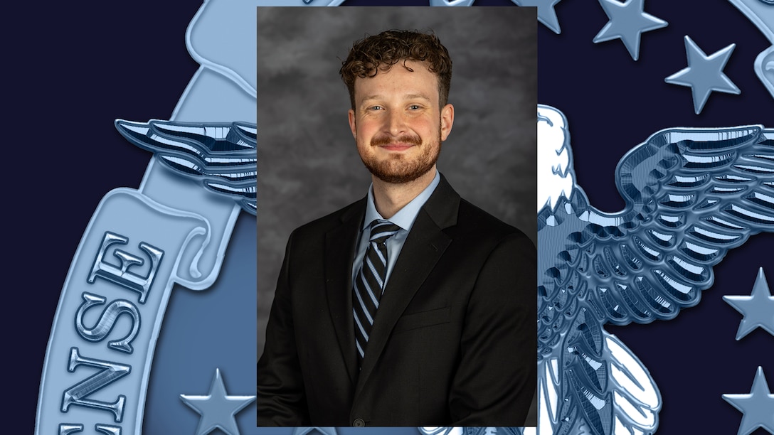 Headshot of man with short, wavy brown hair and short beard and mustache, wearing a black suit with a blue shirt and blue and black diagonal-striped tie.