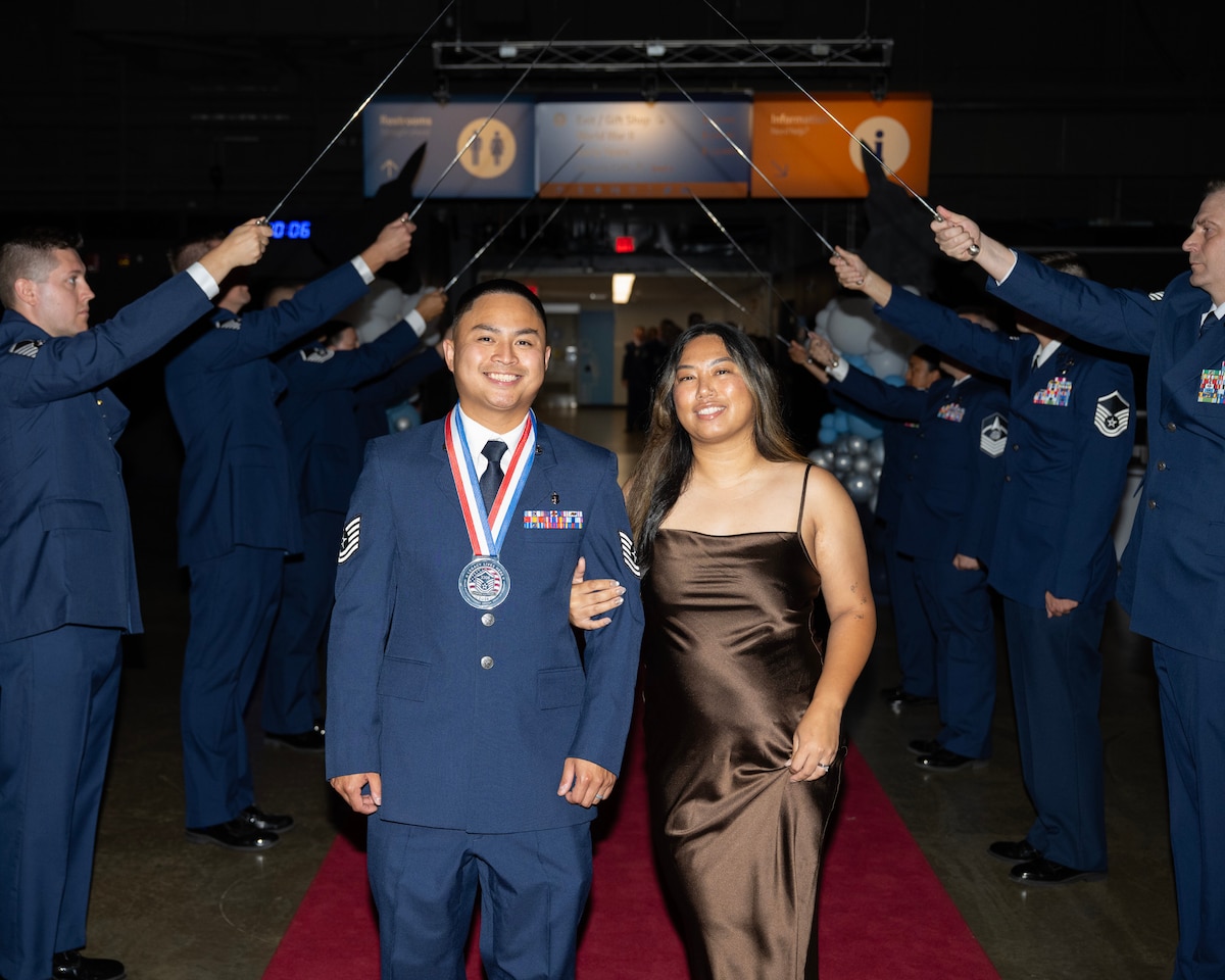 A man in a dress uniform and wearing a medallion stands with his wife, in an evening gown. Behind them men hold swords in the air.