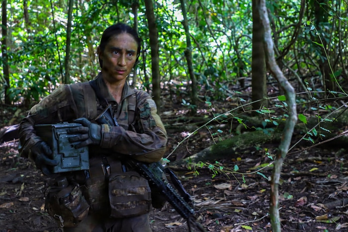 A woman wearing a camouflage military uniform and tactical gear carries an ammo can while walking through the jungle.