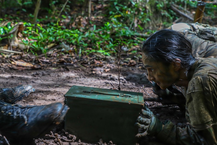 A woman wearing a camouflage military uniform pushes an ammo can through the mud while crawling in the jungle.