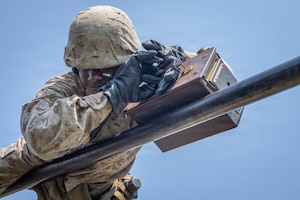A person wearing a camouflage military uniform holds onto an ammo can while moving across an elevated pipe.