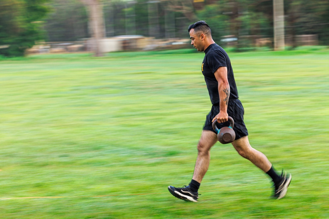 A soldier in a black T-shirt and shorts carries a kettlebell in one hand while moving across a grassy field, with blurry trees in the background.