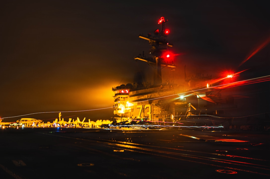 A jet landing on the flight deck of an aircraft carrier in dark conditions creates streaks of light, as red and yellow lights illuminate the control tower in the background.