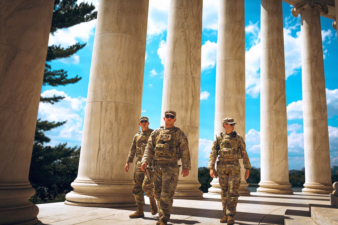 Three guardsmen in camouflage uniforms patrol a monument with large, white marble columns on a bright day, with trees in the background.