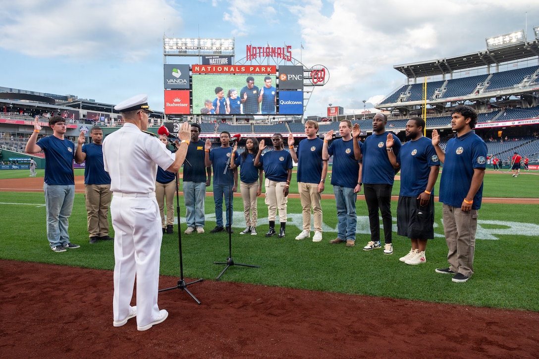 A sailor in white military dress uniform holds up his hand and speaks into a microphone as a group of 13 people wearing dark blue T-shirts face him while holding up their hands on the field in a baseball stadium on a sunny day.